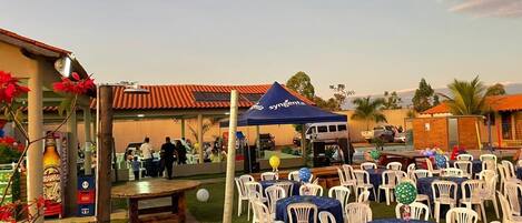 Un área de patio al aire libre con mesas redondas y sillas de plástico blancas, un toldo azul y un enfriador de cerveza.