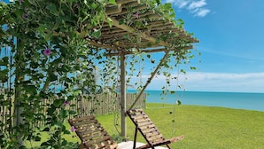 Une pergola en bois, ornée de plantes grimpantes, et deux fauteuils de jardin en bois, installés sur une terrasse donnant sur une pelouse et la mer.