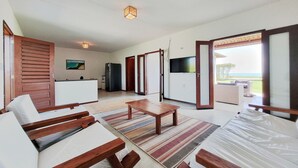 A spacious living area with a wooden sofa, a coffee table, and a TV. There is a kitchen area with a refrigerator and a dining table. The room has large sliding doors leading to a balcony with a view of the outdoors. - Beach Front Villas - House by the sea by Carpediem (Aracati)