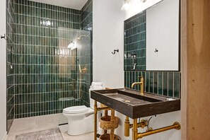 Casa de banho com chuveiro em cabine de vidro, sanita branca, lavatório escuro com torneira dourada e espelho sobre o lavatório.