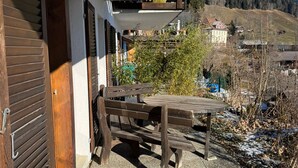En uteplats med ett träbord och bänkar, en dörrmatta och utsikt över ett berg och hus i bakgrunden.