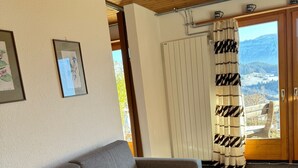 Ett mysigt vardagsrum med en grå soffa, två mönstrade kuddar och ett fönster med utsikt över ett bergigt landskap.