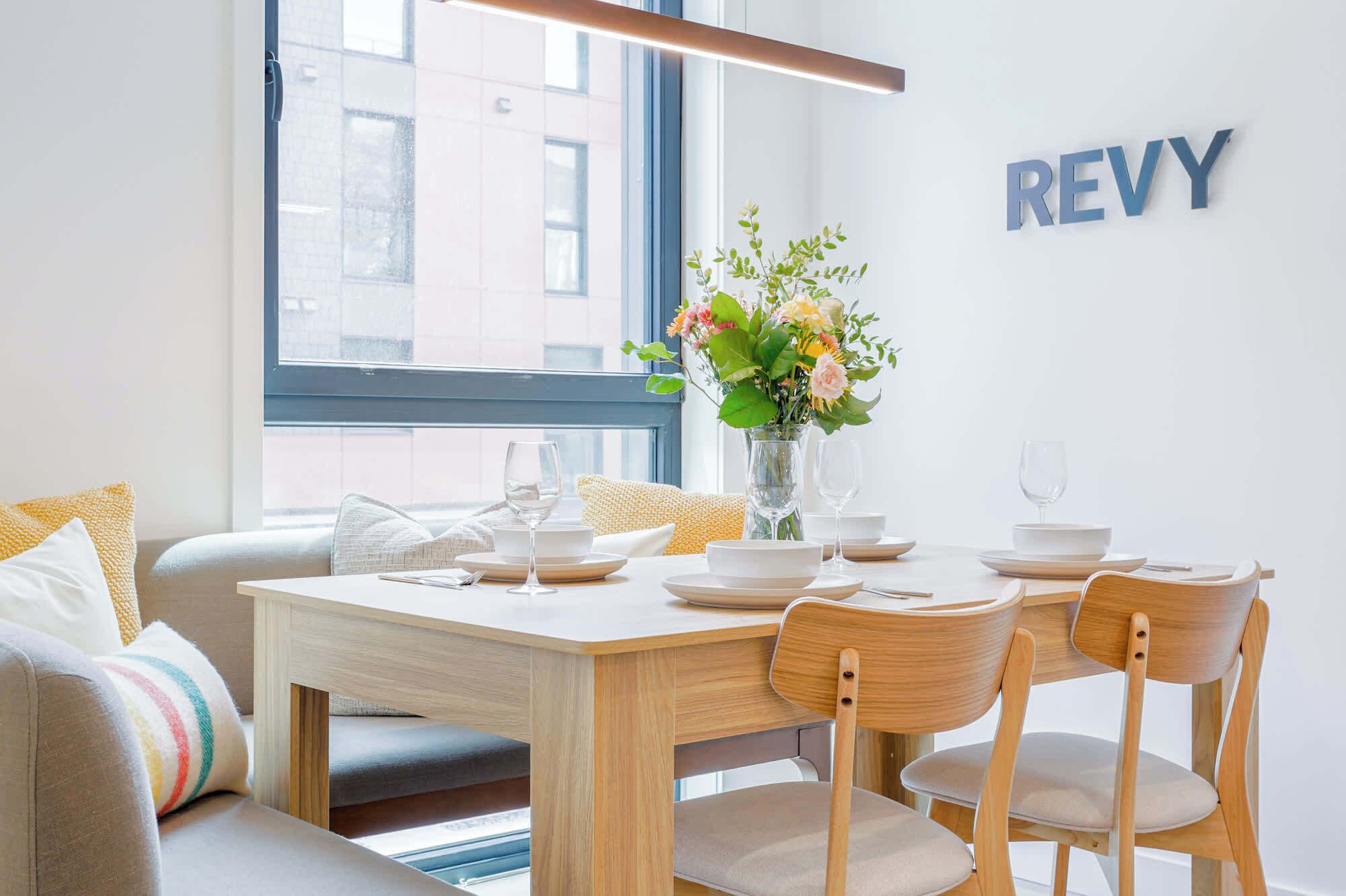 A dining area with a wooden table set for two, a vase with flowers, and a 'REVY' sign on the wall.