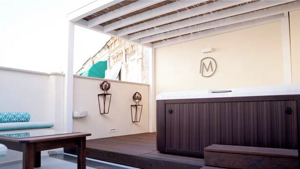 Uma área de pátio com mesa de madeira, um banco e uma banheira de hidromassagem. Há arandelas decorativas e um logotipo na parede.