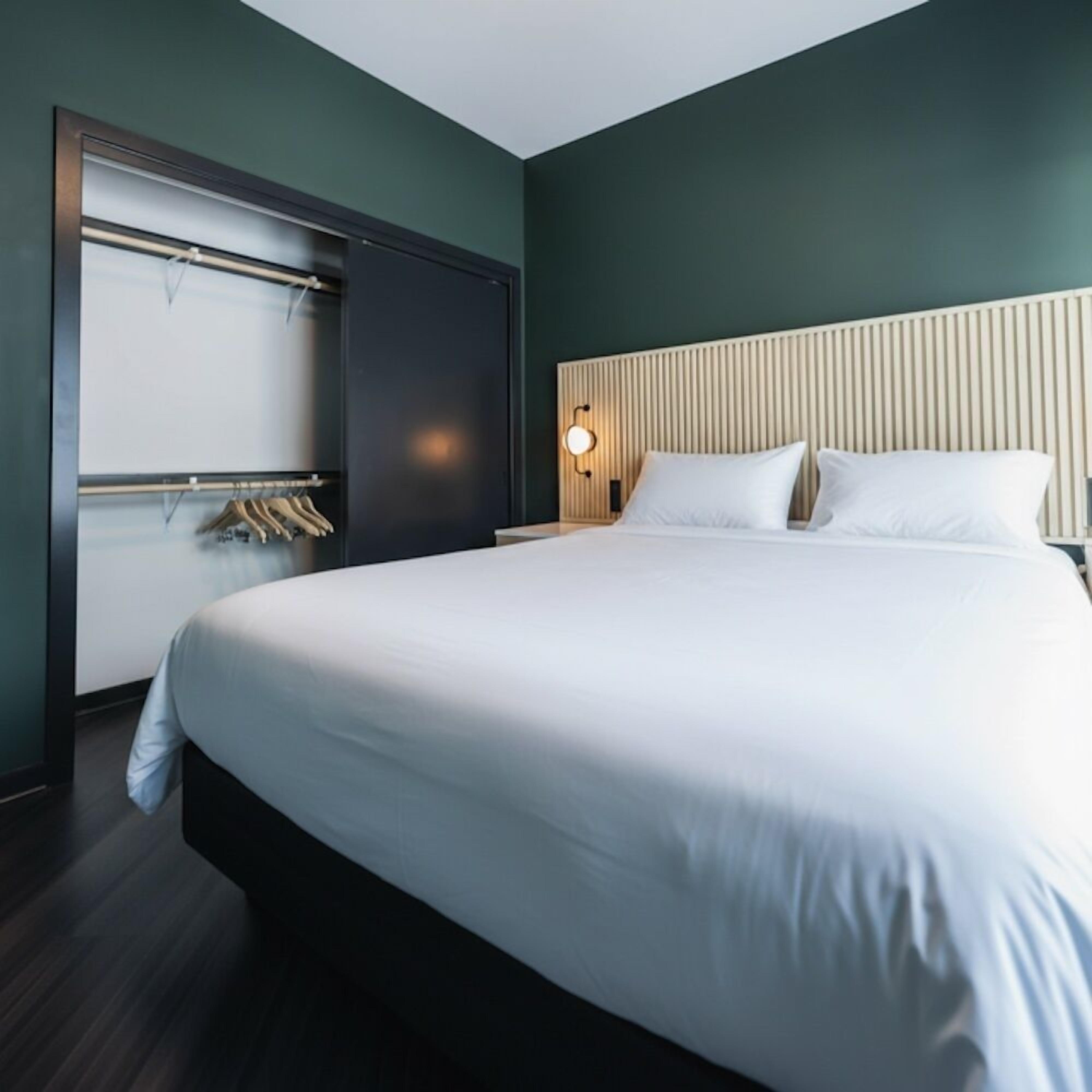 A modern hotel room with a large bed, a wardrobe, and a bedside table with a lamp.