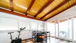 Ein Zimmer mit Schachbrettmuster-Boden, einem Tisch mit weißer Spitzenleinentuch und einer Blume, Ergometer, ein Esstisch mit Stühlen und große Fenster mit Jalousien.