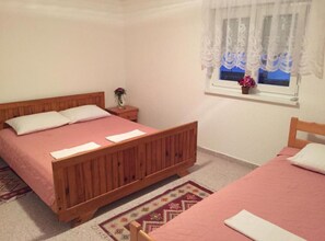 A bedroom with two beds, a nightstand with a vase of flowers, a window with lace curtains, and a patterned rug. - Apartments Bozena - Two bedroom apartment (Krk)