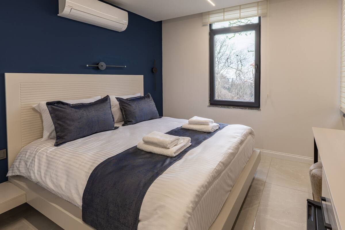 A bedroom with a large bed, a bedside table, a window with a view of trees, and an air conditioning unit.