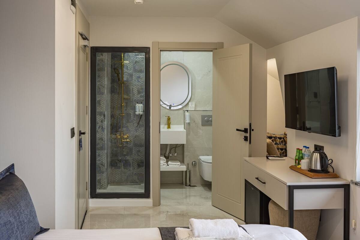 A modern hotel room with a glass-enclosed shower, a large mirror, a flat-screen TV, and a bedside table with toiletries.