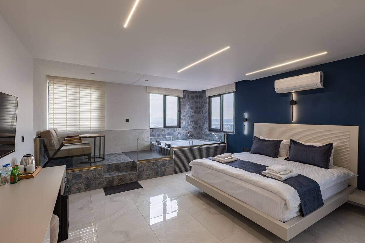A modern bedroom with a large bed, a desk, a seating area, and a bathtub.