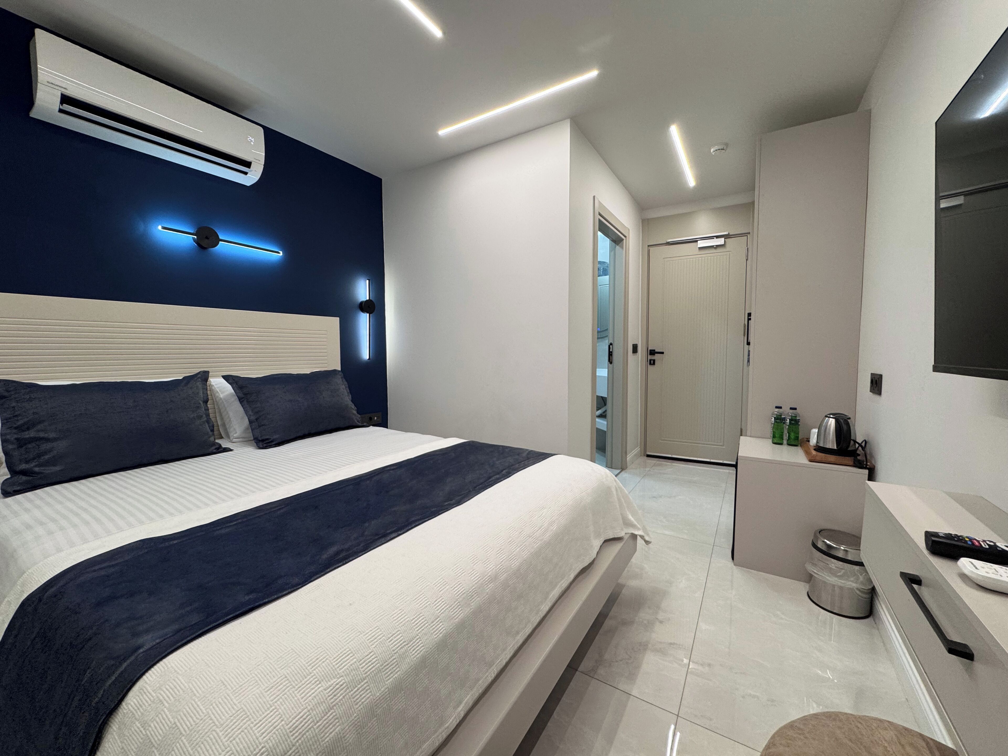A modern hotel room with a large bed, bedside tables, an air conditioning unit, a mirror, and a door leading to a bathroom.