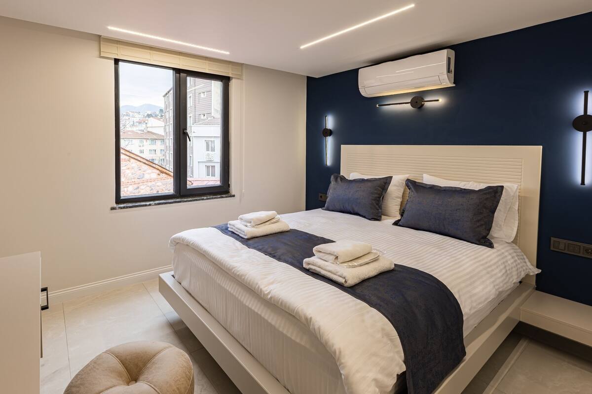 A modern bedroom with a large bed, a window, an air conditioner, and a bench.