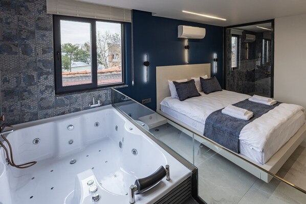 A modern bedroom with a large bathtub, a bed with a headboard, and a window with a view of a building. - Safir Termal&Spa Otel (Bursa)