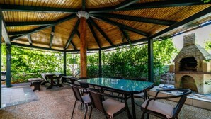 Une terrasse extérieure couverte, dotée d’une table et de chaises à plateau en verre, et entourée de verdure.