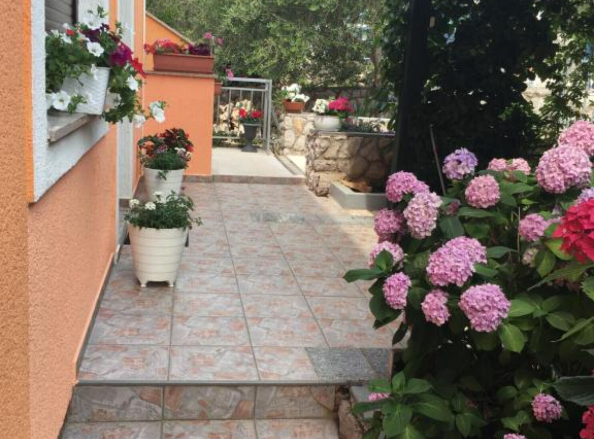 Ein schmaler Pfad, gesäumt von Kübelpflanzen und Blumen, der zu einer Steinmauer und einem Garten führt.