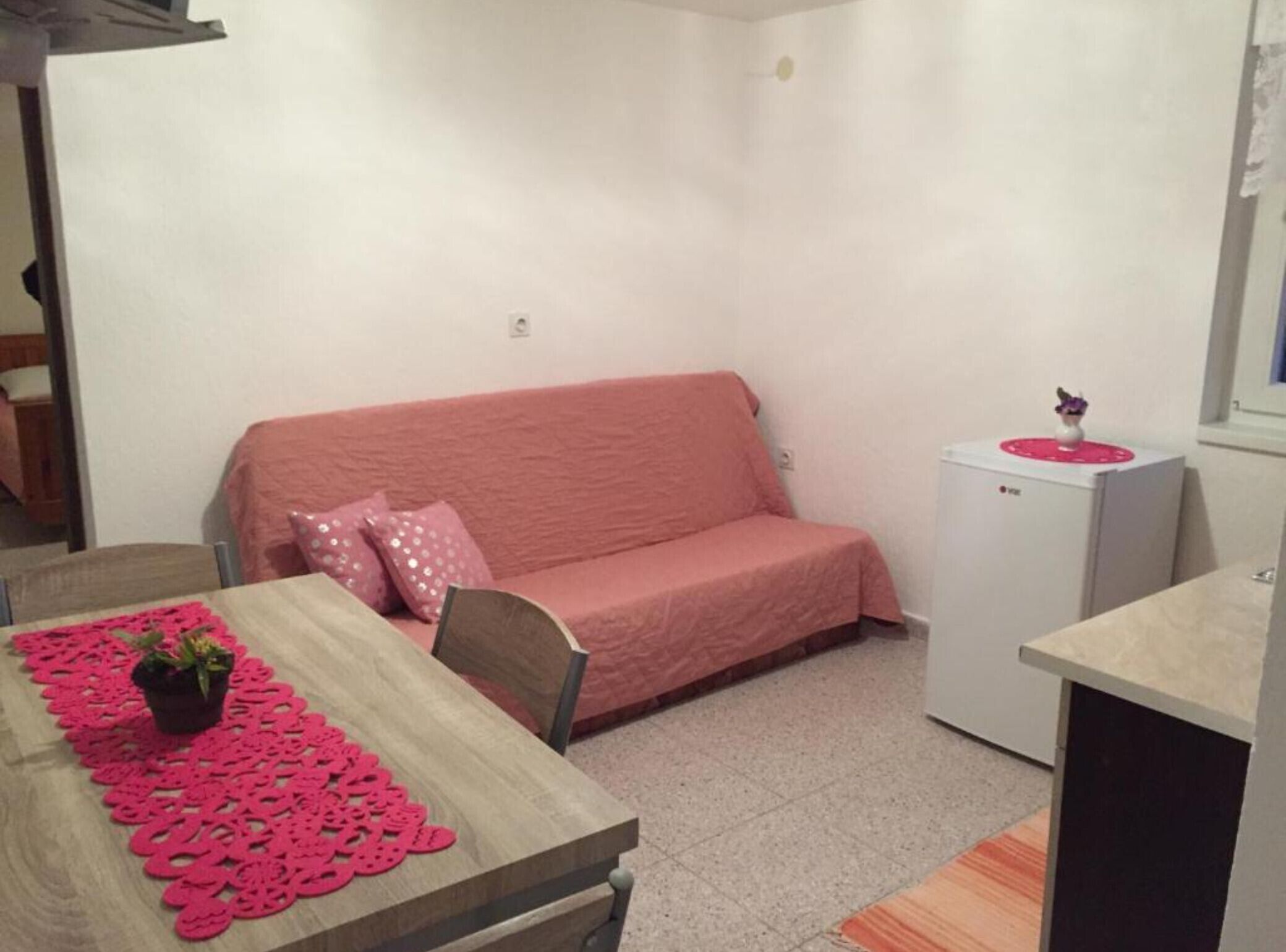 Um quarto pequeno e aconchegante com um sofá rosa, uma mesa de madeira com um jogo americano rosa e uma geladeira branca.