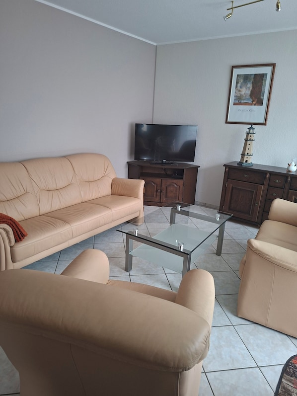 Un salon avec un canapé beige, une table basse en verre, un téléviseur posé sur un meuble en bois et un tableau encadré accroché au mur.
