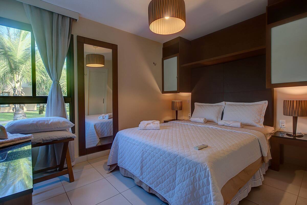 A well-lit bedroom with a large bed, bedside tables, a mirror, and a view of palm trees outside.