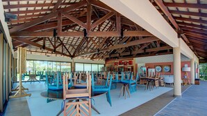A spacious indoor area with multiple tables and chairs, a high ceiling with exposed beams, and a view of the outdoors through large windows. - VG Sun #102 - Apto de Dois Quartos por Carpediem (Caucaia)