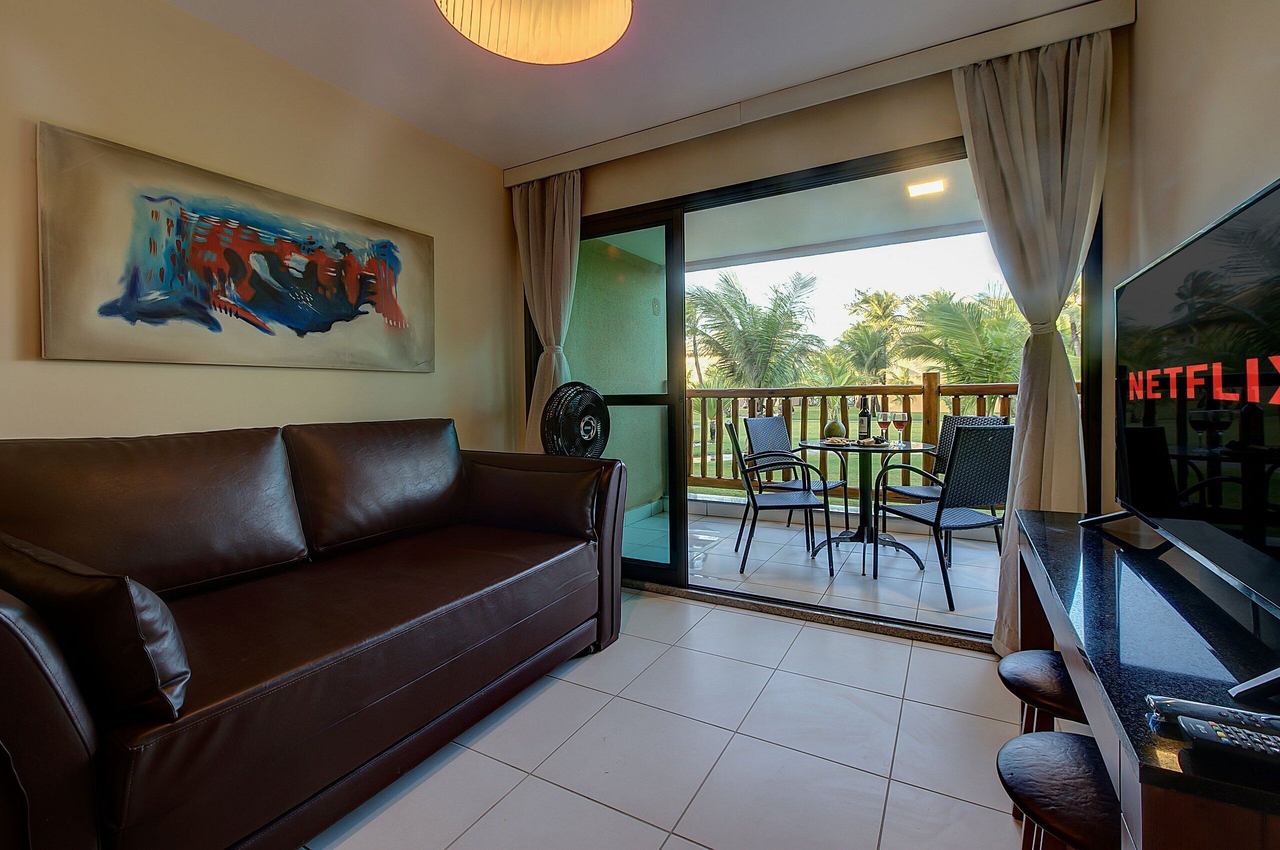 A living room with a brown leather sofa, a flat-screen TV, a painting on the wall, and a balcony with chairs and a table.