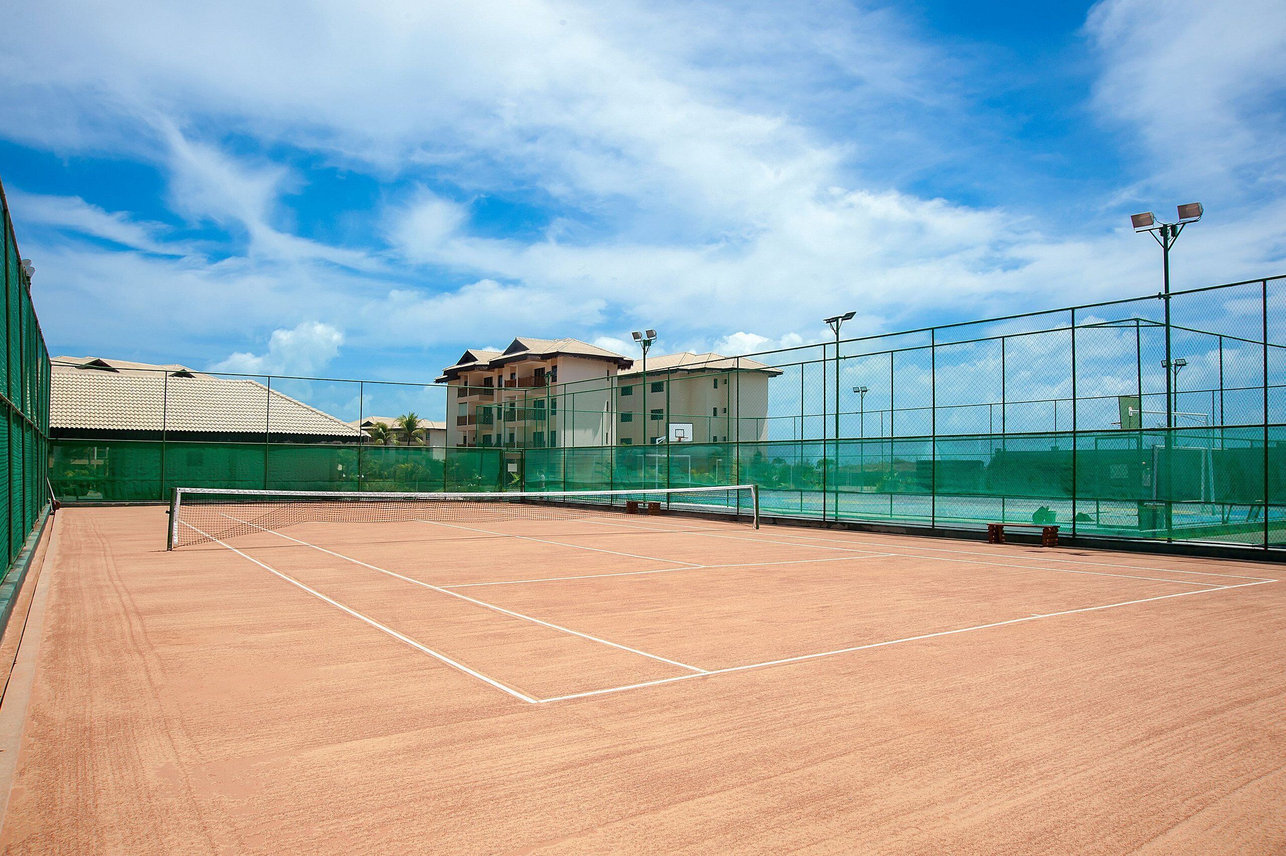 Una cancha de tenis al aire libre con una red, rodeada por una cerca verde y un edificio al fondo.