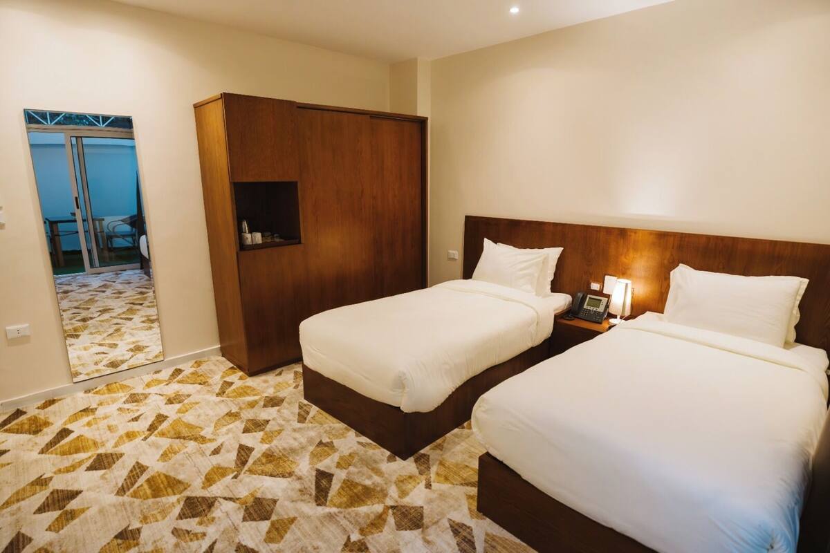 A hotel room with two beds, a wooden wardrobe, a patterned floor, and a view of the outside.