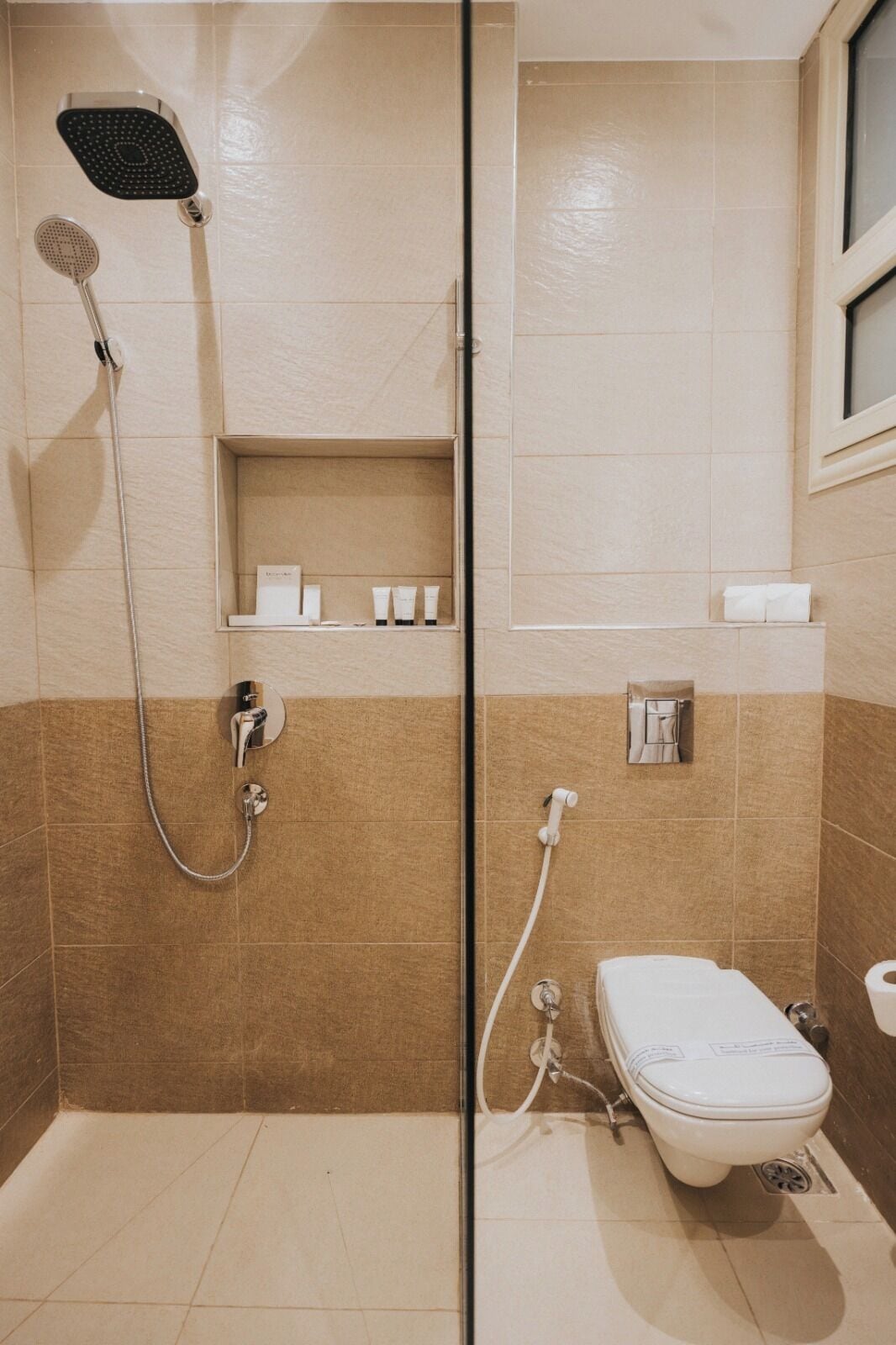 Un baño con mampara de vidrio, cabezal de ducha, inodoro y estante con artículos de aseo.