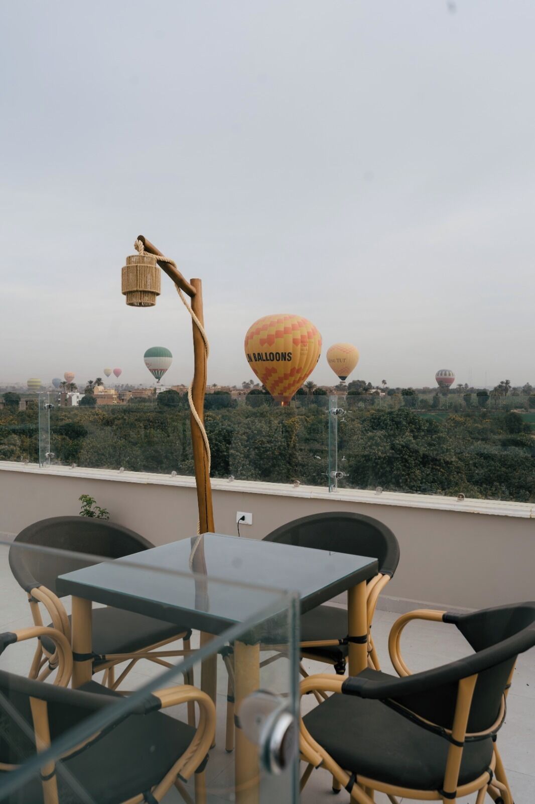 Un balcón con sillas de mimbre y una mesa de vidrio, con vistas a un paisaje urbano donde se divisan globos aerostáticos a lo lejos.