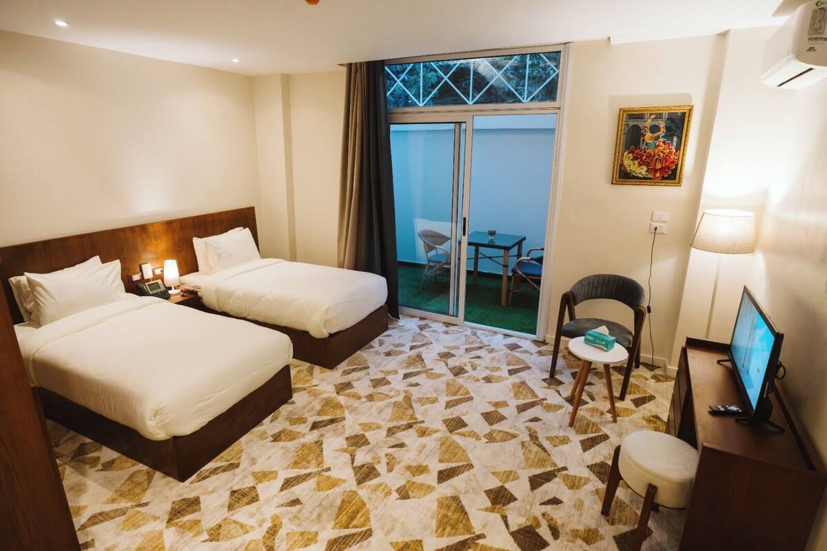 A hotel room with two beds, a desk with a computer, a chair, a TV, and a balcony with a table and chairs.