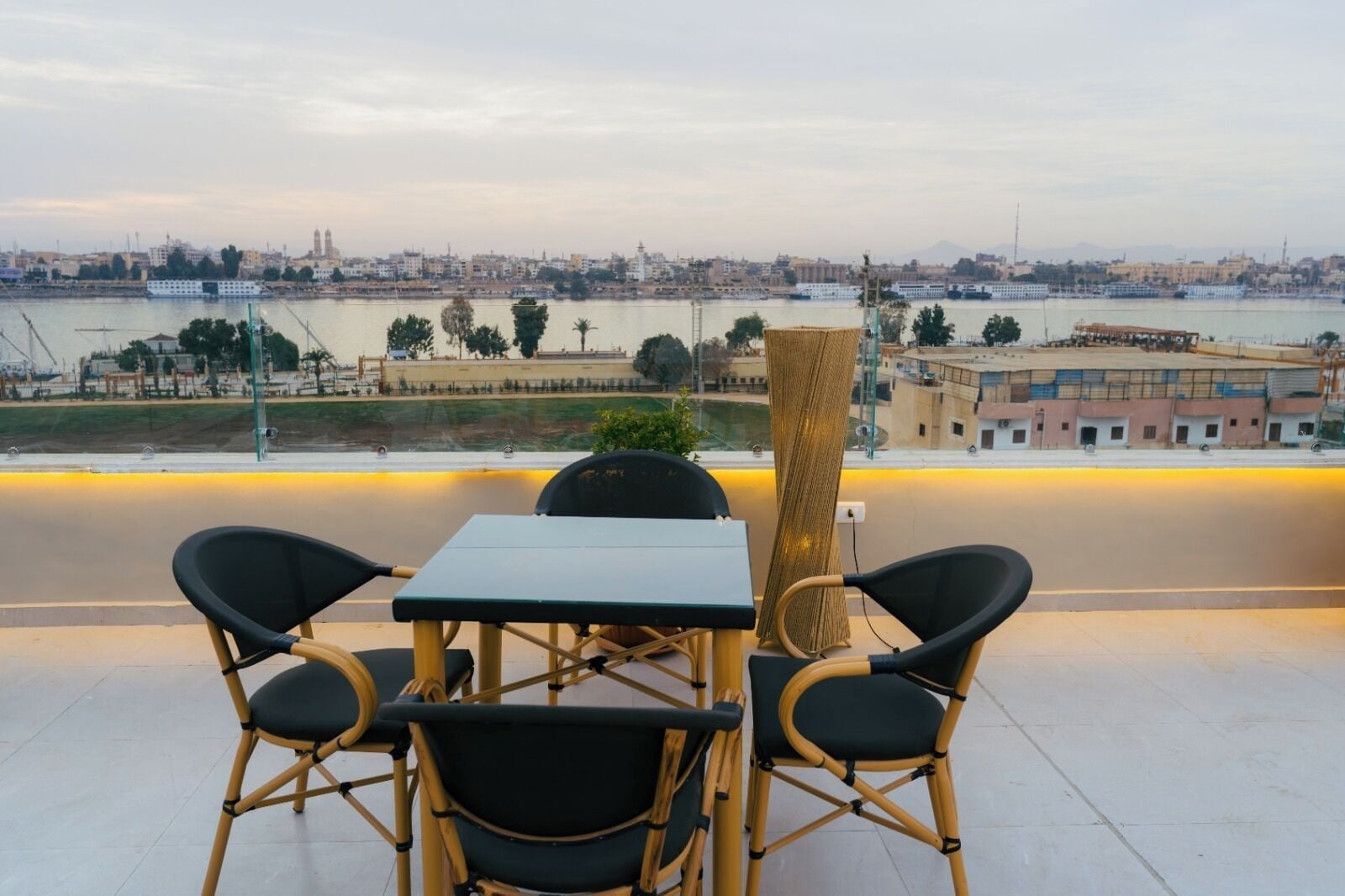 A terrace with a table and chairs overlooking a river with boats, buildings, and a cityscape in the background.