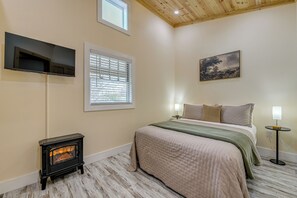 A bedroom with a bed, a wall-mounted TV, a window with blinds, a painting, a small table with a lamp, and a wood-burning stove. - Walk to Main St! Mtn-View Getaway in Waynesville (Waynesville)