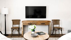 A modern living room with a flat-screen TV mounted on the wall, a wooden coffee table with a vase of flowers, two wicker chairs, and a floor lamp. - Auburn Elite Retreat (Opelika)