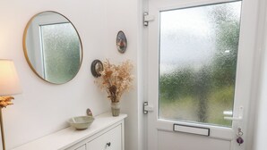 A white door with frosted glass, a white wall with a round mirror, a small vase with dried flowers, and a bowl on a white cabinet. - Pump Cottage (Truro)