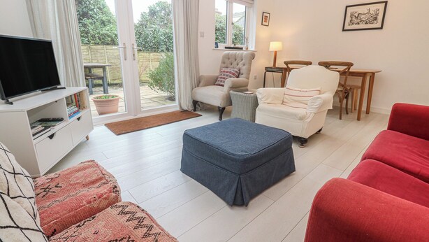 A living room with a TV, two armchairs, a red sofa, a small table, and a window with a view of greenery. - Pump Cottage (Truro)