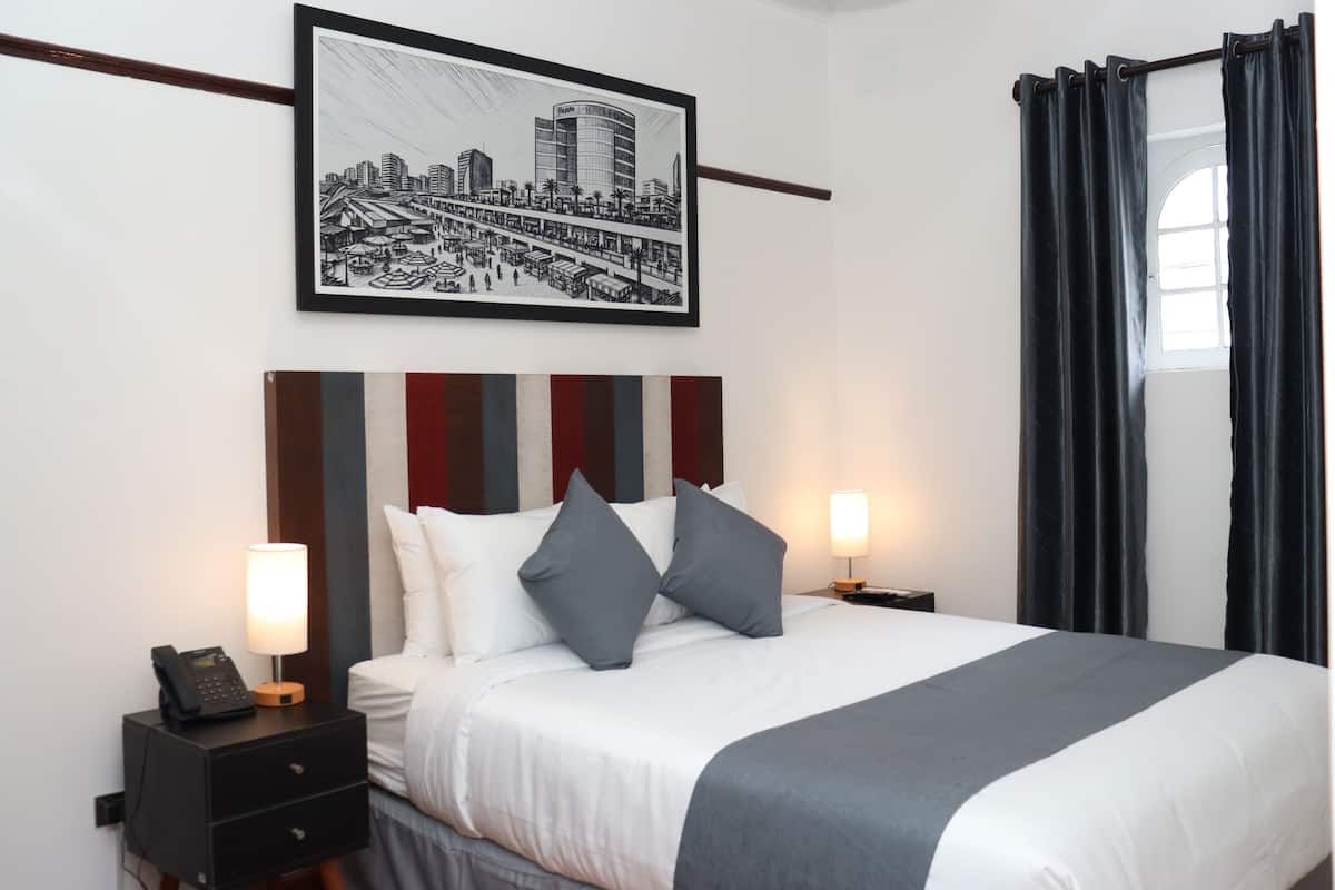 Habitación de hotel con cama, dos mesitas de noche, una lámpara de noche, una obra de arte con un paisaje urbano enmarcado y una ventana con cortinas.