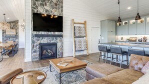 A spacious living area with a stone fireplace, a large flat-screen TV, and a kitchen with bar stools. - 'Scenic Overlook' - Luxe Greers Ferry Hideaway! (Fairfield Bay)