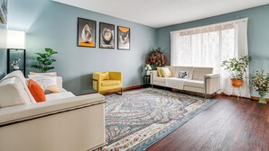 Una sala de estar con un sofá color beige, un sillón amarillo, un sofá blanco, un tapete estampado y un ventanal con cortinas translúcidas.