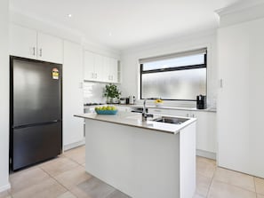 A modern kitchen with white cabinetry, a stainless steel refrigerator, a central island with a bowl of fruit, and a window above the sink. - Azura on Gordon Mornington Townhouse Near Beach (Mornington)