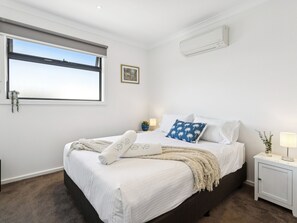 A bedroom with a large bed, a window with a view, a bedside table with a lamp, and an air conditioning unit on the wall. - Azura on Gordon Mornington Townhouse Near Beach (Mornington)