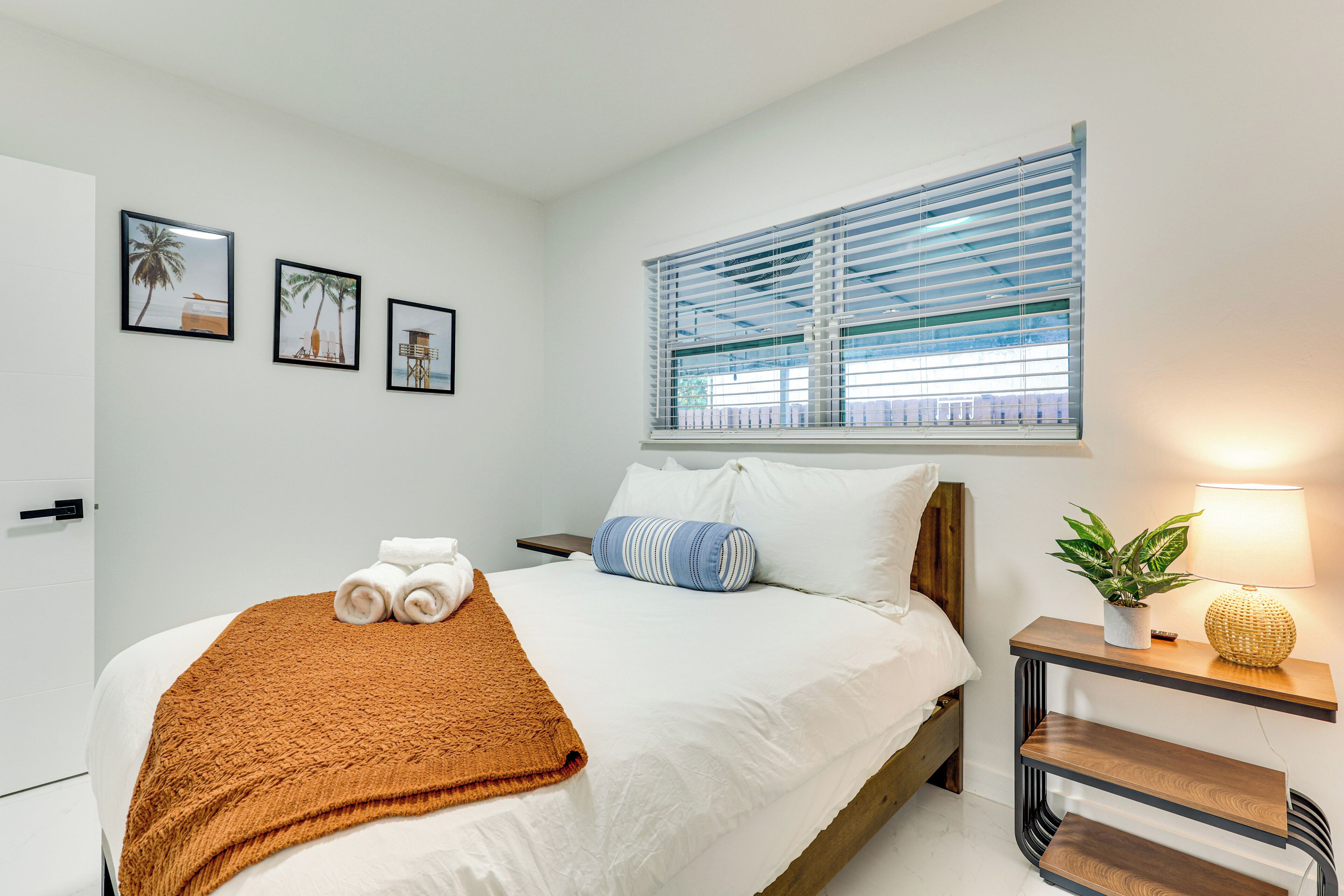 Un lit bien fait, recouvert d’une couverture marron, avec des oreillers blancs et un coussin à rayures bleues. Trois tableaux encadrés ornent le mur, il y a une fenêtre avec des stores, et une table de chevet sur laquelle se trouvent une lampe et une plante.