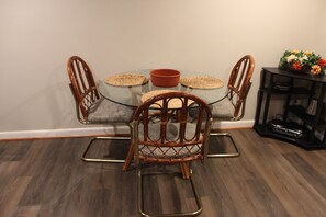 A dining area with a glass table, four wicker chairs, a red bowl, and a basket of flowers on a side table. - Cozy Eclectic Lanham Suite - Close to DC! (Lanham)
