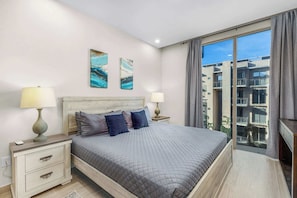 A modern bedroom with a large bed, two bedside tables, a nightstand, and a view of a cityscape through floor-to-ceiling windows. - Las Arenas 1BR | Pool, Jacuzzi, Near Beach (Cabo San Lucas)