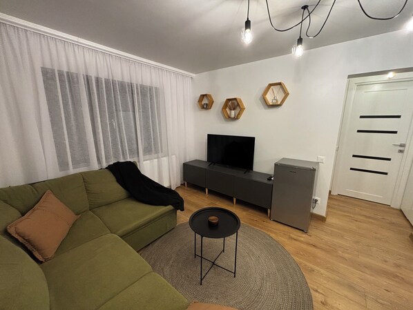 Una sala de estar moderna con un sofá verde, un televisor de pantalla plana, una mesita y un refrigerador.