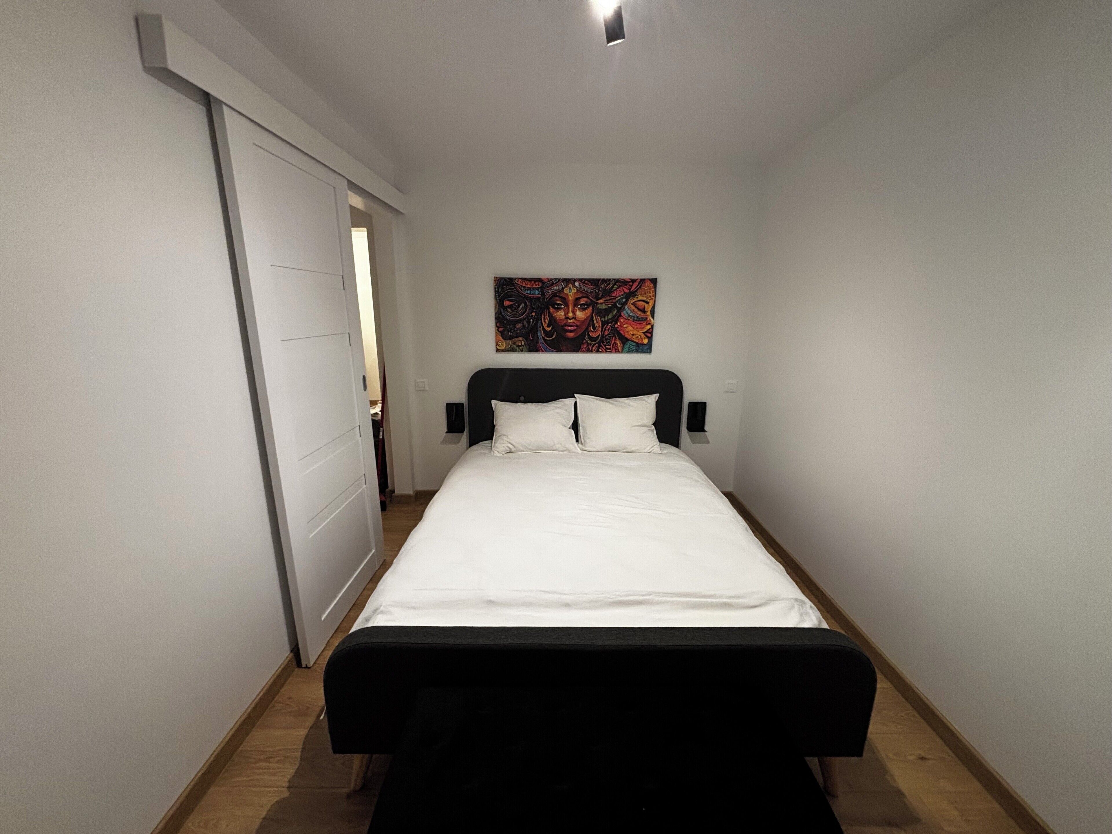 A narrow hotel room with a bed, a door, and a painting on the wall.