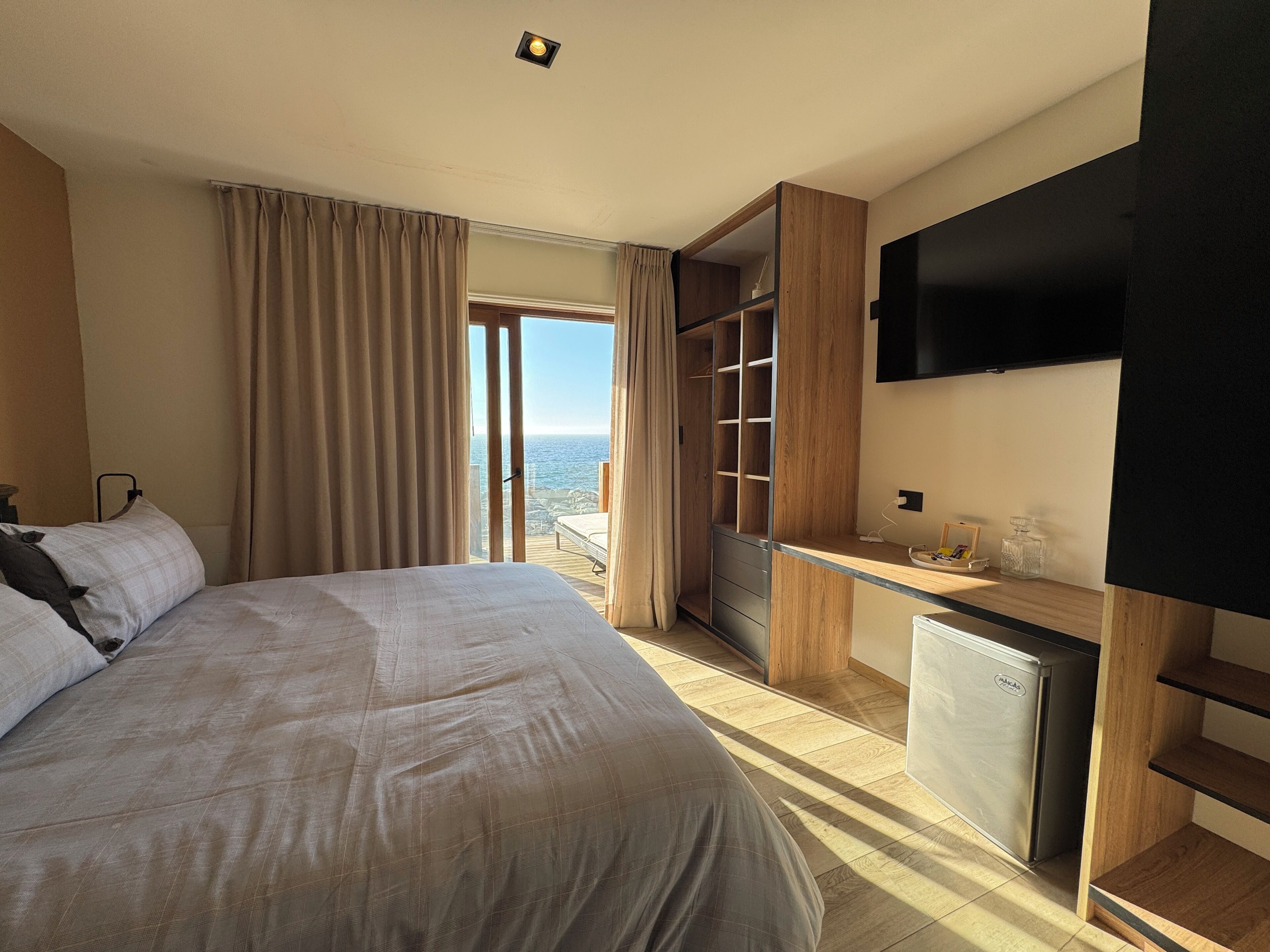 A hotel room with a large bed, a desk with a refrigerator, a TV, and a view of the ocean.