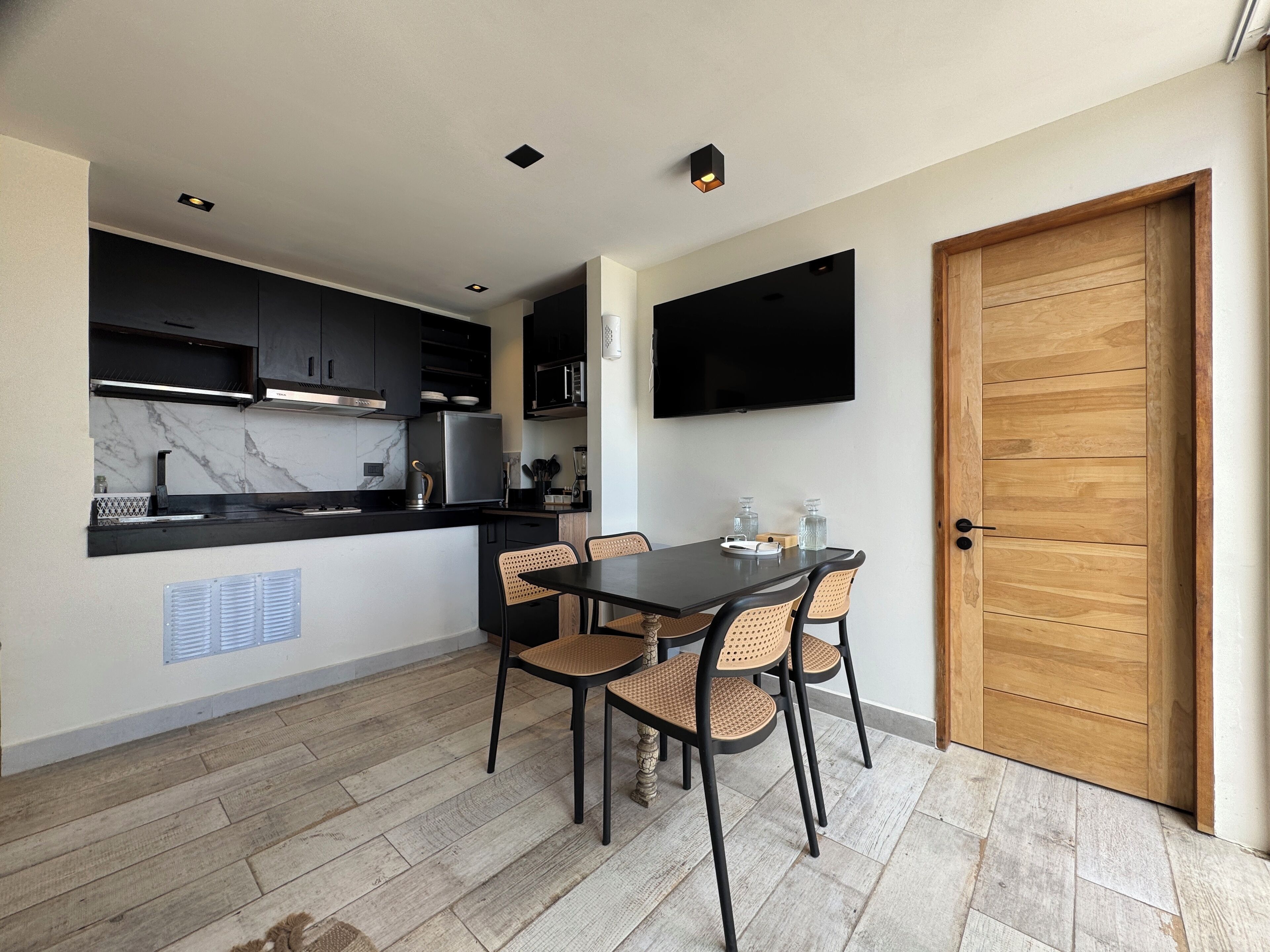 A compact kitchen with modern appliances, a dining area with wicker chairs, and a flat-screen TV mounted on the wall.
