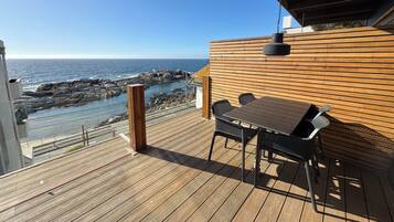Una terraza de madera con mesa y sillas con vistas a la costa rocosa y al océano.