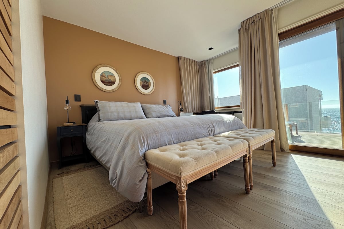 Un dormitorio con una cama grande, un banco, una mesita de noche, dos lámparas de pared y un ventanal con cortinas.