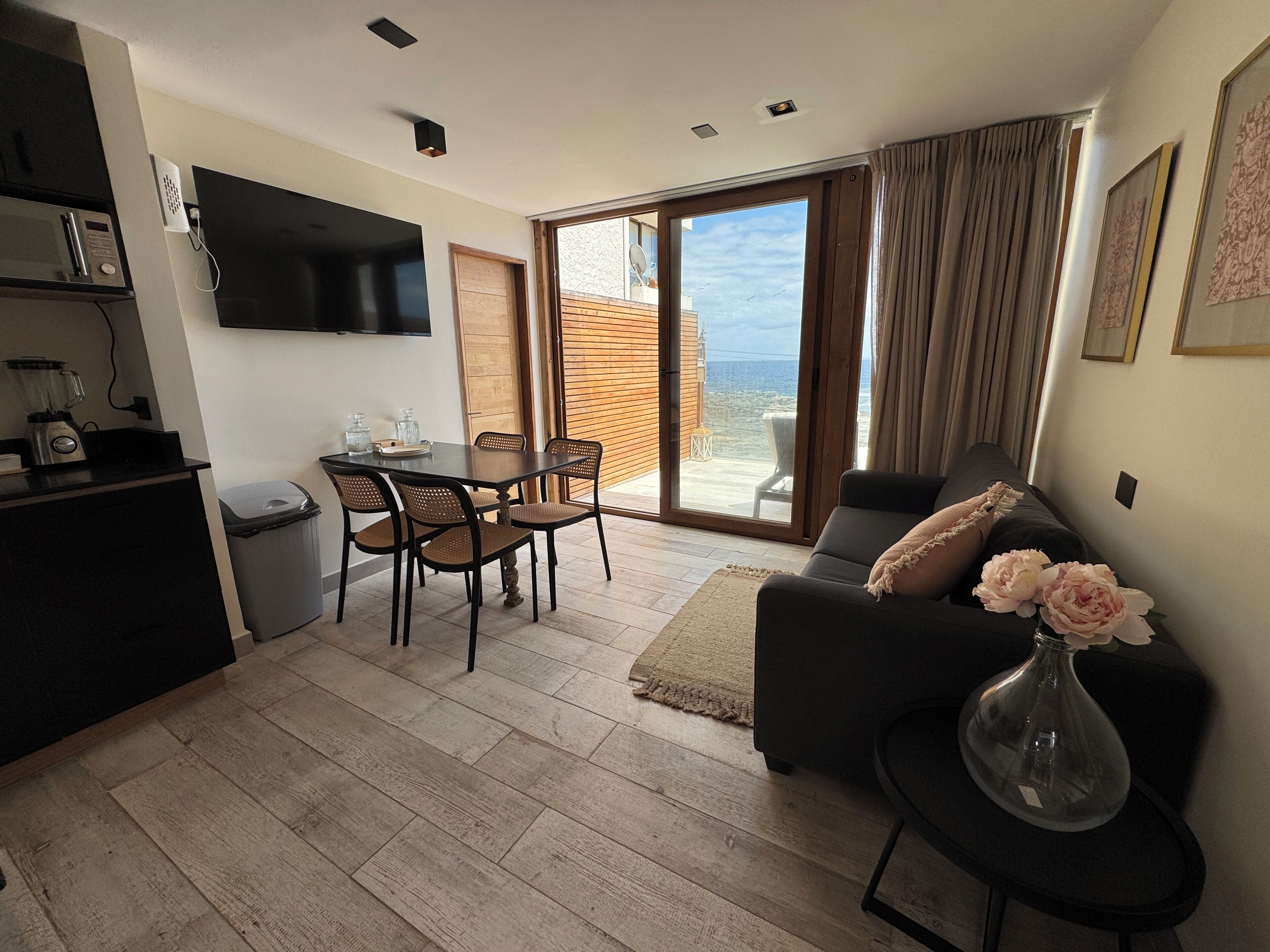 A modern living room with a sofa, a dining table with chairs, a TV mounted on the wall, and a sliding glass door leading to an outdoor area.