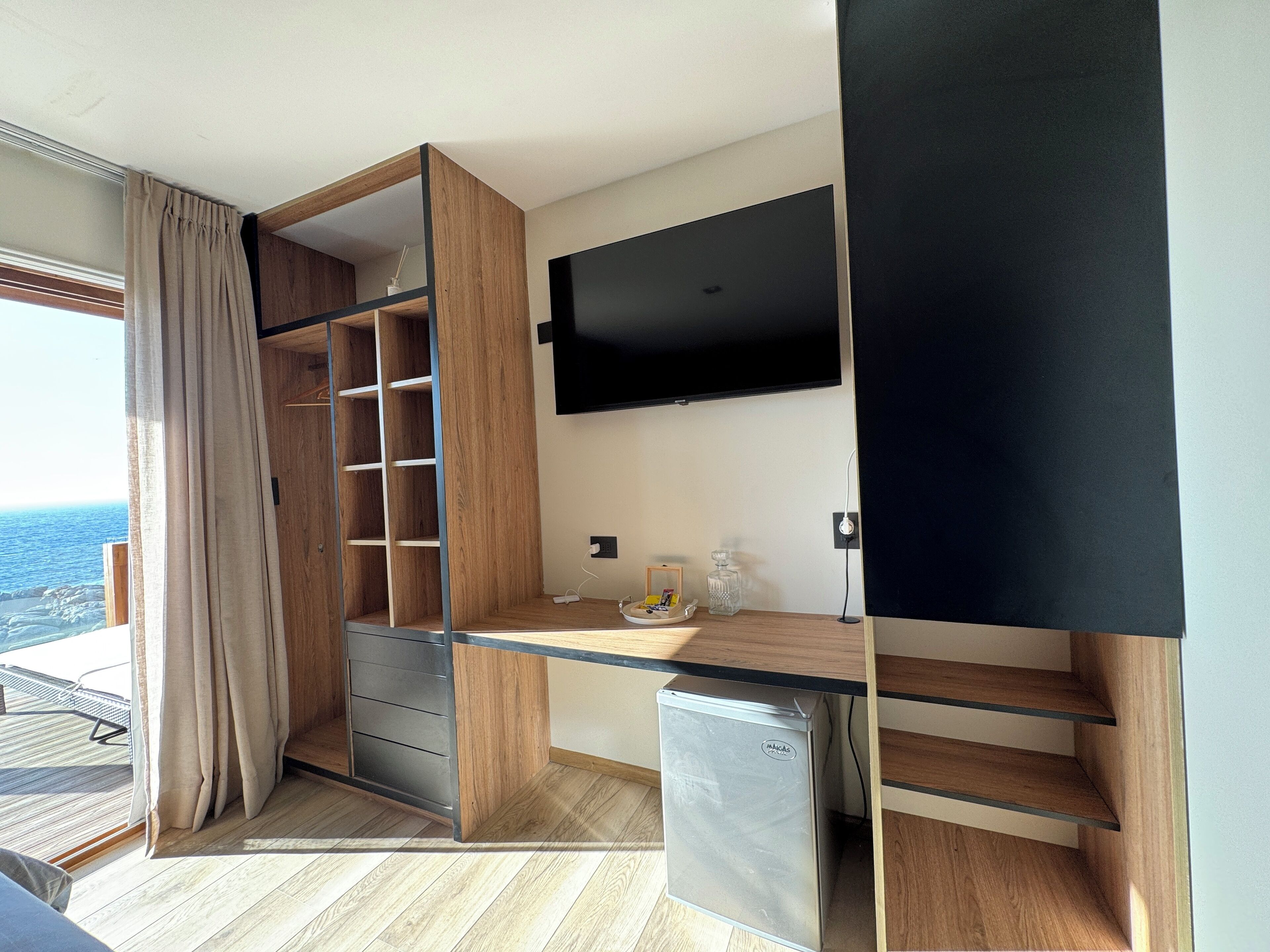 A compact room with a built-in desk, a flat-screen TV, a mini-fridge, and a sliding wardrobe with a view of the sea.